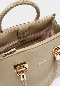 Sac à main en cuir beige avec une poignée tressée, des ferrures dorées, une fermeture éclair et un intérieur rose pâle avec une poche. Logo visible à l'intérieur.