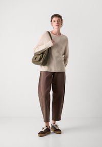 Kvinna klädd i beige tröja, bruna byxor, bruna sneakers och bärande en olivgrön axelväska, stående mot en vit bakgrund.