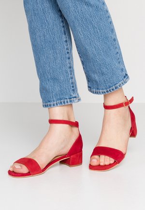 Fødder i røde ruskindsandaler med lav blokhæl og ankelremme, kombineret med cropped blå denimjeans mod en ensfarvet baggrund.