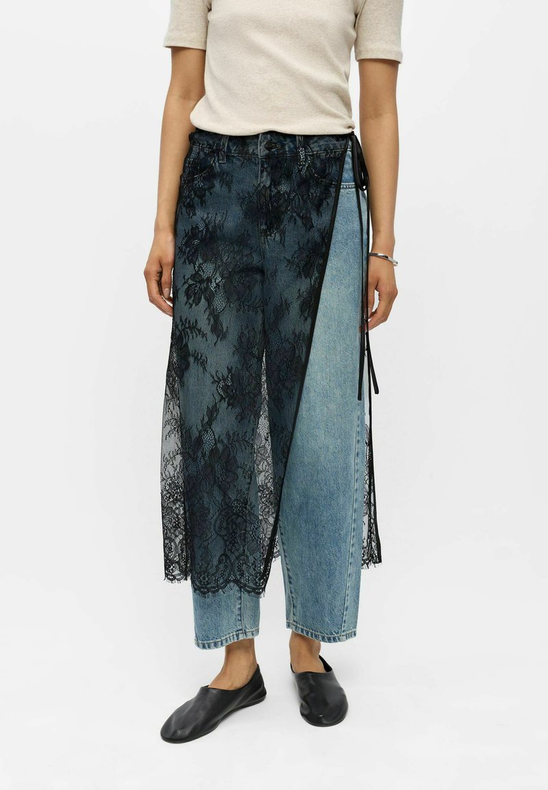 Femme portant un jean bleu avec un overlay en dentelle noire, un haut beige à manches courtes, des chaussures noires slip-on, debout sur fond blanc.