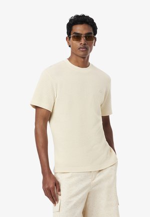 Uomo con camicia beige texturizzata a maniche corte e pantaloncini abbinati, occhiali da sole, in piedi davanti a uno sfondo bianco uniforme.