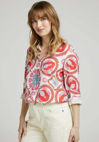 Chemise à col avec des motifs floraux et feuillus colorés, présentant des nuances de rouge, bleu et rose sur un fond clair ; manches longues retroussées.