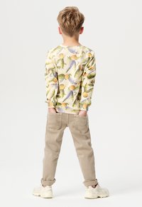 Lichtgele overhemd met lange mouwen met fruit- en bladmotieven, gecombineerd met beige broek. Het kind staat met de handen in de zakken en draagt witte sneakers.