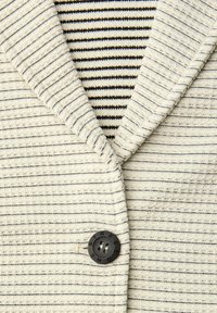 Crèmekleurig vest met dunne zwarte horizontale strepen, een donkere knoop met het label "STREET ONE", over een zwart-wit gestreept shirt.