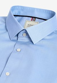 Hellblaues Hemd aus einem strukturierten Stoff mit einem festlichen Kragen und zwei weißen Knöpfen. Mit einem Label in Rot und Schwarz.