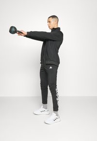Black athletic jacket and pants, white logo on pants, gray socks, and white shoes. Holding a kettlebell, with a straight posture.