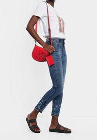 Bolso bandolera rojo con diseño moteado en negro, camiseta blanca con estampado decorativo, vaqueros azules con bordado de paisley, sandalias negras de pala.