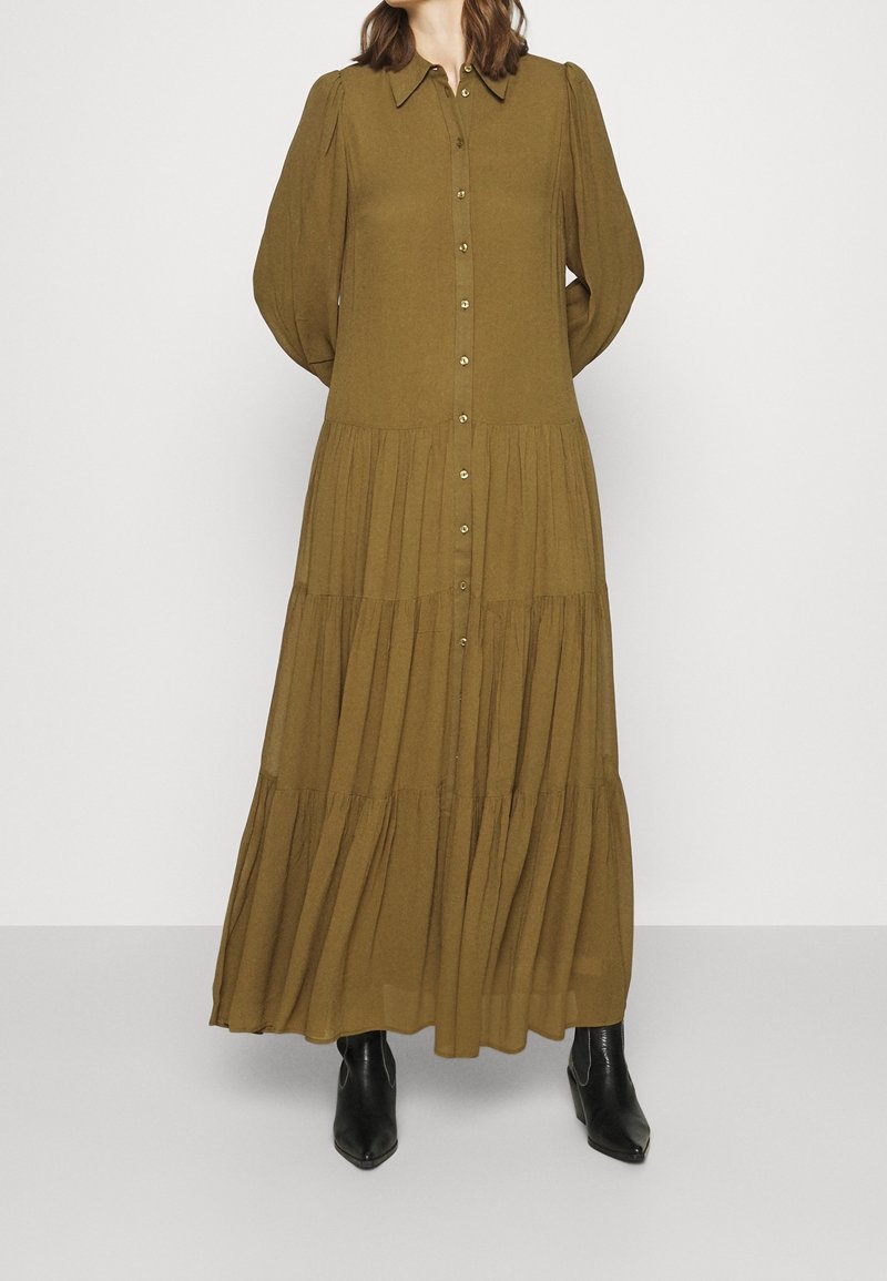 Femme portant une longue robe olive à volants avec des boutons sur le devant et des bottines noires à talons, debout devant un fond uni.