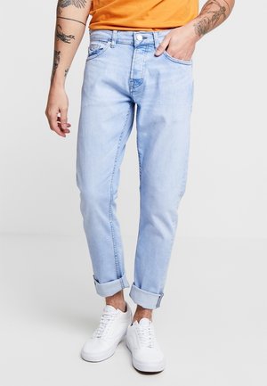 Personne portant un jean bleu clair retroussé, des baskets blanches et un t-shirt orange. Tatouages visibles sur les deux bras. Debout devant un fond uni.