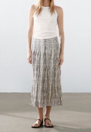 Vrouw draagt een witte mouwloze top, een geplooide midi rok met slangenprint en zwarte sandalen met zilveren accenten, staand op een effen vloer.