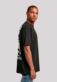 Schwarzes Baumwoll-T-Shirt mit kurzen Ärmeln, Rundhalsausschnitt und großem weißen Text sowie grafischem Design auf der Rückseite. Seitenansicht mit hellblauen Jeans.