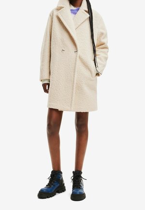Abrigo beige de gran tamaño con textura, solapa tipo notch y doble botón, acompañado de botas de cordones rugosas en azul y negro.