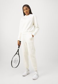 Felpa bianca e pantaloni sportivi realizzati in morbido tessuto, con un piccolo logo rosa. La modella tiene in mano una racchetta da tennis nera e indossa scarpe bianche.