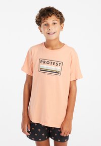 Lichtoranje katoenen T-shirt met een rechthoekige graphic met daarop "PROTEST" en strepen. Gecombineerd met zwarte shorts met een palmbomenpatroon.