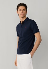 Mężczyzna o ciemnych włosach, ubrany w granatową koszulkę polo z krótkim rękawem i białe spodnie, stojący na jednolitym jasnoszarym tle.