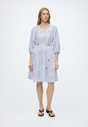 Frau trägt ein weiß-blaues, knielanges Kleid mit Blumenmuster, 3/4-Ärmeln und einem dünnen Seilgürtel, kombiniert mit schwarzen Sandalen mit Absatz, steht still.