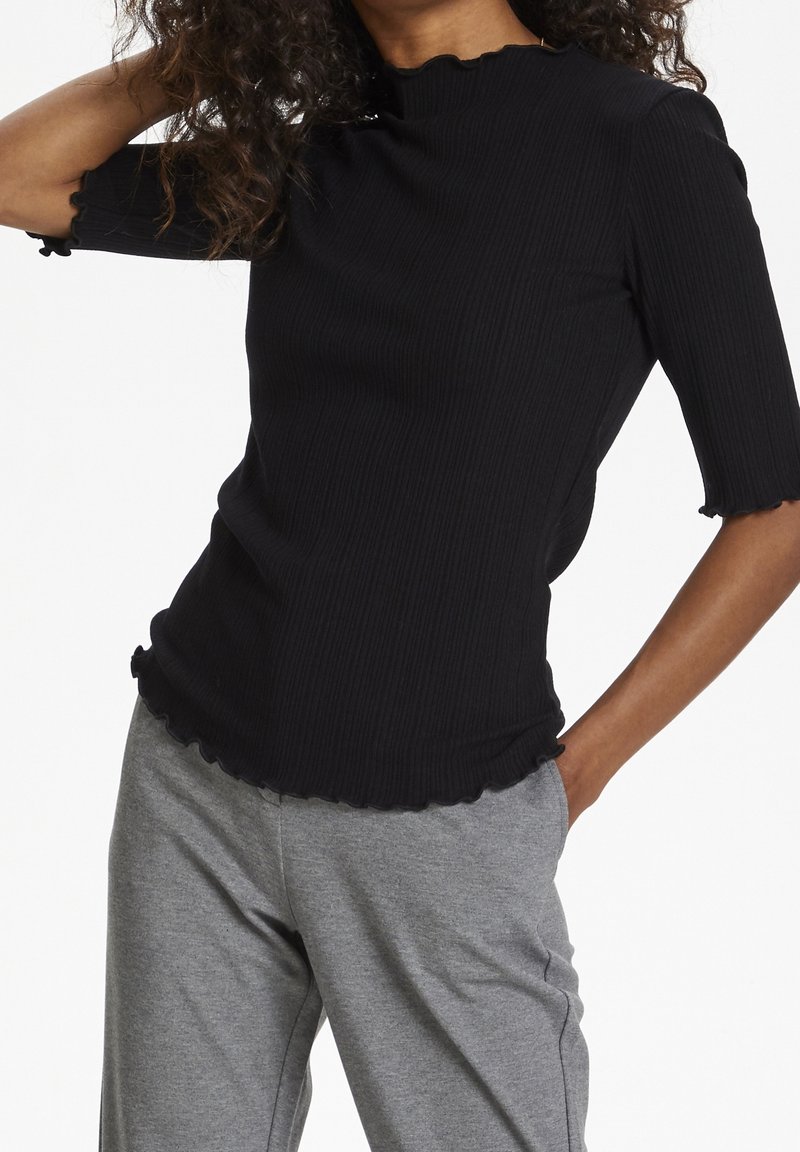 Kobieta w czarnym, prążkowanym topie z krótkim rękawem i falbankami na krawędziach oraz szarych casualowych spodniach, pozująca z jedną ręką w kieszeni.