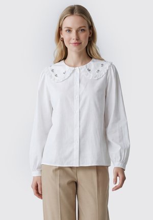 Ung kvinde med langt blondt hår iført en hvid knapbluse med en bølget krave og beige bukser, står foran en ensfarvet baggrund.