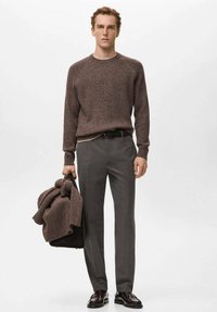 Jersey marrón acanalado con cuello redondo, pantalones grises a medida y zapatos oscuros pulidos. El modelo sostiene una chaqueta marrón a juego.