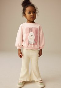 Rosa Pullover mit einem Hasenmotiv und gepunkteten Ohren; kombiniert mit cremefarbenen Schlaghosen und Schuhen mit grünen Akzenten. Weiche Texturen sind sichtbar.