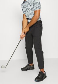 Černé golfové kalhoty, světle modrá vzorovaná polo košile a černé golfové boty. Golfista drží hůl a soustředí se na úder.