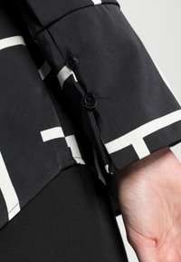 Blusa negra con patrones geométricos blancos, que cuenta con un puño con dos botones negros y una textura suave. Primer plano de la zona de la manga.