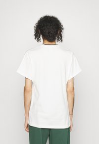 Vit bomullströja med korta ärmar, rak skärning och en svart detalj vid halsringningen, sedd bakifrån, kombinerad med gröna byxor.