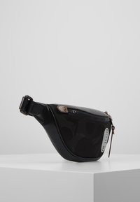 Černá kožená a textilní ledvinka se zipovým uzávěrem, kulatým tvarem, strukturovaným povrchem a malým kovovým zdobením na přední straně.