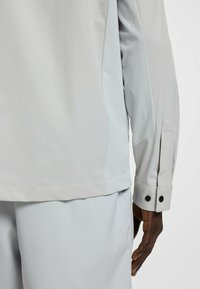 Light gray long-sleeve shirt with textured side panels, flat seams, and buttoned cuffs. Soft fabric and relaxed fit extend to the waist.