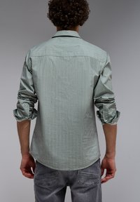 Raidallinen paita vaaleanvihreänä ja valkoisena, pitkät hihat kainalossa, kaarevalla helman muotoilulla. Pehmeä kangas, jossa sileä rakenne. Takakatsaus.