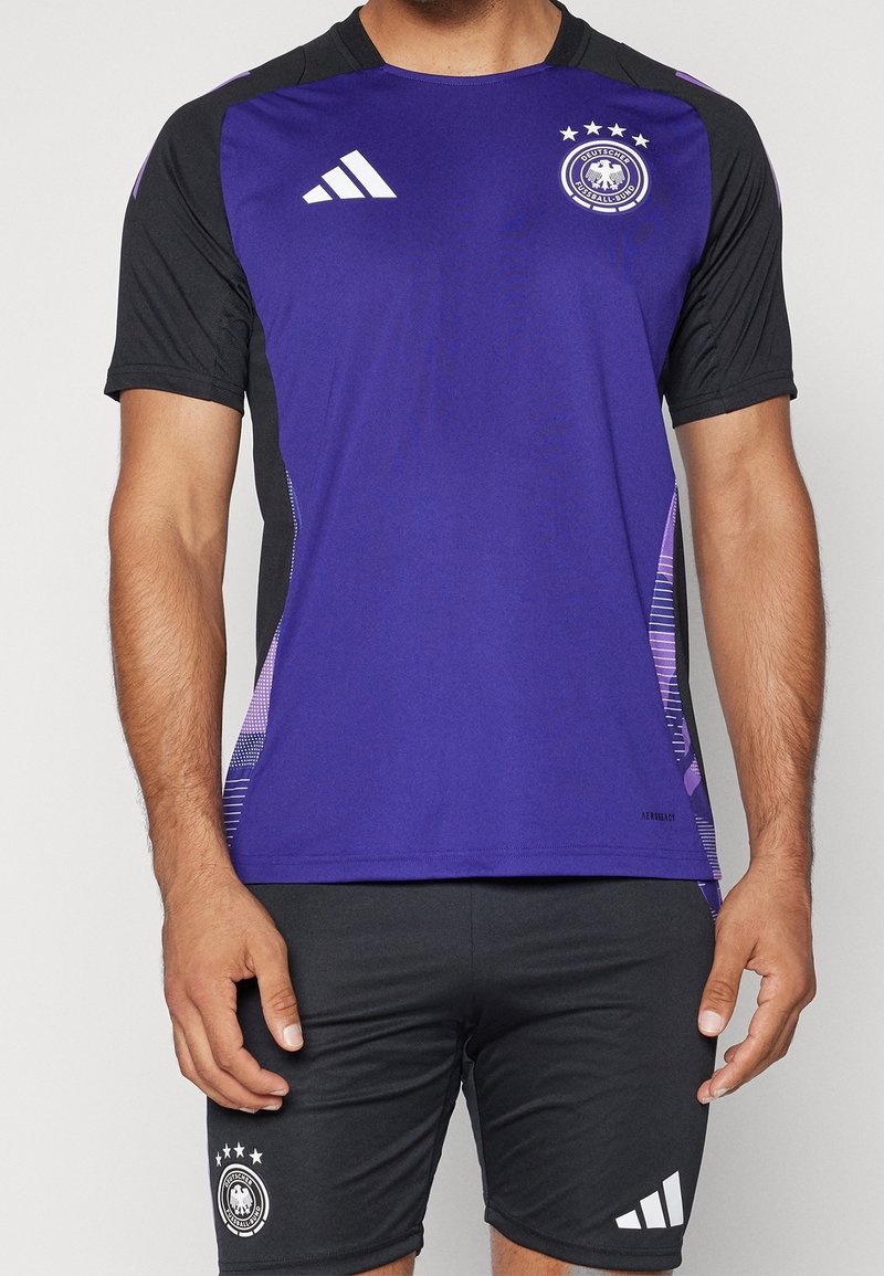 Lila och svart sporttröja med rund hals, korta ärmar och tre vita ränder på axeln; inkluderar en logotyp och grafiska detaljer.