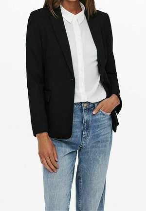 Frau trägt einen schwarzen Blazer über einem weißen Hemd mit Knöpfen sowie hellblaue Jeans, eine Hand in der Tasche, beschnittener Ansichtsbereich des Mittelabschnitts.