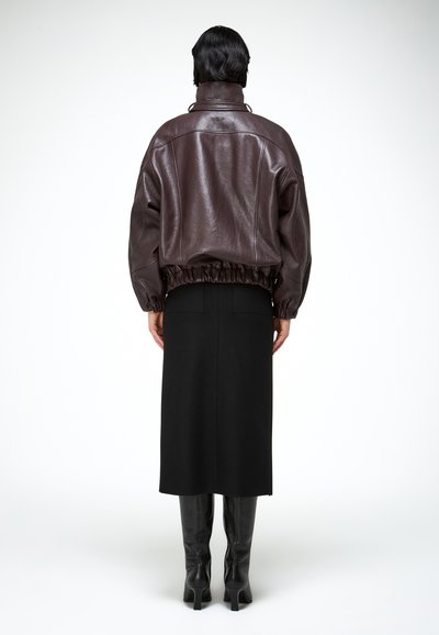 Osoba v tmavo hnedej koženej bunde s elastickým lemom, čiernej midi sukni a čiernych čižmách na podpätku po kolená, otočená chrbtom na bielom pozadí.