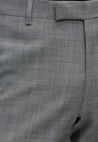 Primo piano di pantaloni in tessuto grigio strutturato con passante per cintura e tasca laterale visibile.