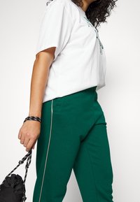 Valkoinen puuvillapaita, jossa on painettu kuvio, yhdistettynä vihreisiin polyesteriseen juoksuhousuihin, joissa on sivusauma. Musta nyöriolosäkki kädessä.