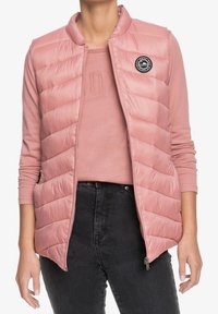 Lichtroze gewatteerd bodywarmer met doorgestikt ontwerp, ritssluiting en zijzakken. Draagt over een bijpassend shirt met lange mouwen en zwarte broek.