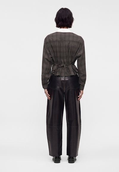 Personne aux cheveux courts et foncés portant un haut marron à carreaux à manches longues noué à la taille et un pantalon en cuir noir à jambes larges, debout de dos.