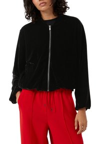 Zwarte fluwelen bomberjack met een rits aan de voorkant, elastische manchetten en zoom; gestyled met rode wijde broek. Heeft een zijzak.