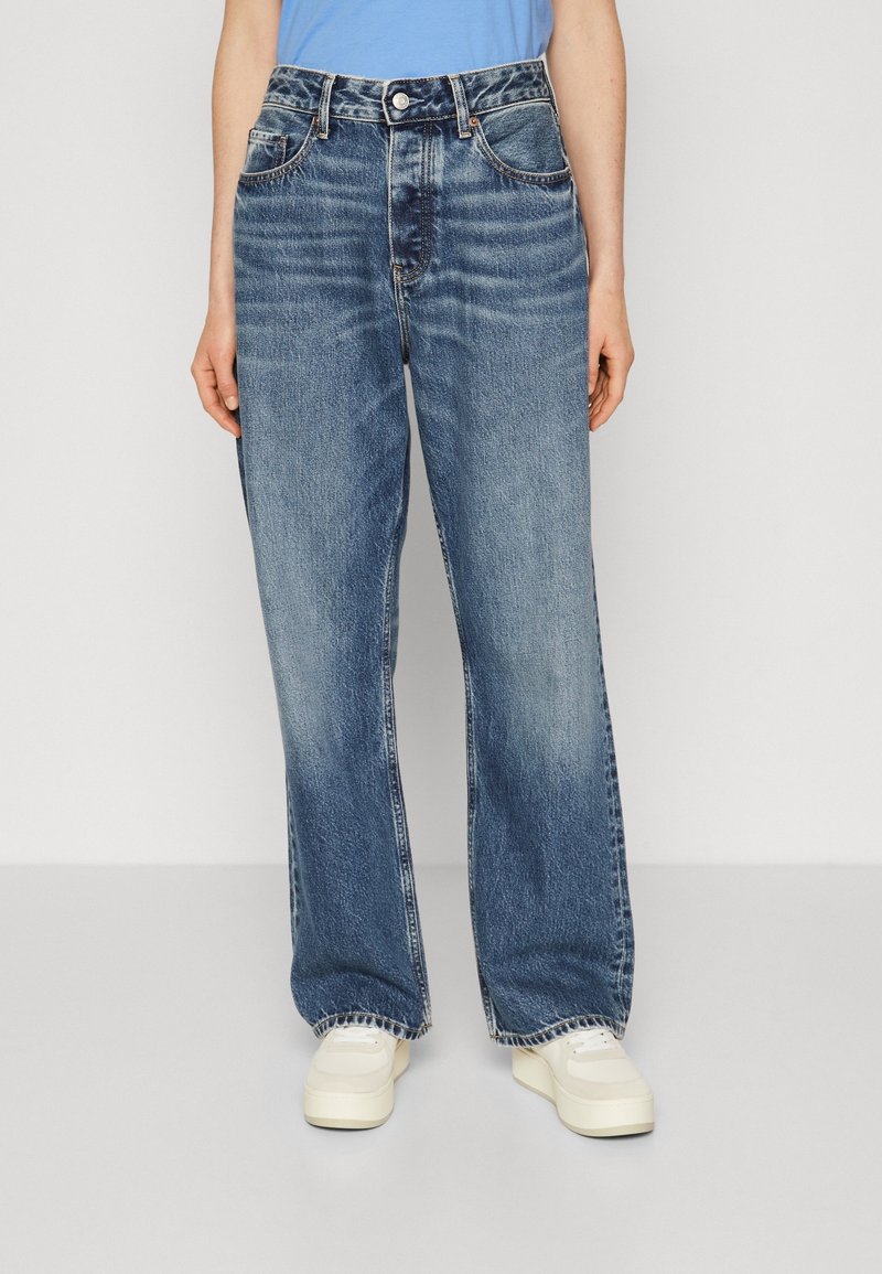 Kvinnor som bär vida blå denimjeans och vita sneakers, står mot en enfärgad ljus bakgrund.