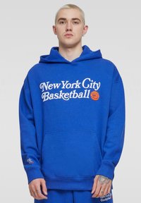 Niebieska bluza z kapturem z miękkiego materiału, z białym napisem "Basketball w Nowym Jorku" oraz grafiką piłki koszykowej, posiadająca kieszeń z przodu i ściągacze u rękawów.