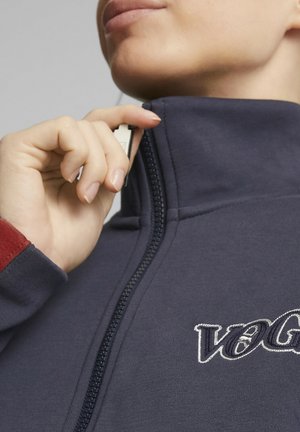Hand pulling up zipper on navy blue jacket with "VOG" embroidered on chest, wearing a garment with red cuff detail.