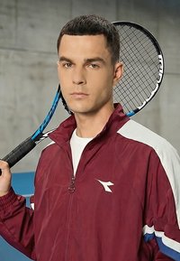 Tânăr purtând o geacă sportivă bordo cu alb, ținând o rachetă de tenis pe umăr, stând în interior.