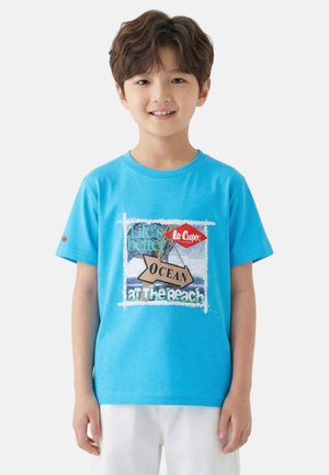 Garçon souriant portant un T-shirt bleu avec un motif de plage océanique et un short blanc sur un fond blanc uni.
