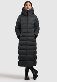 Sort pufjakke med høj krave, der har lynlås foran, horisontalt quiltet design og sidelommer, parret med knæhøje sorte støvler.