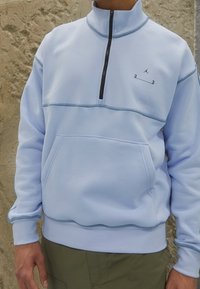 Světle modrý svetr s čtvrtzipem a klokankou, s černým zipem, jemnými švy a logem na hrudi.