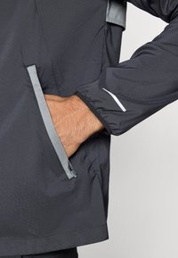 Σκούρο, ελαφρύ μπουφάν με υφή. Διαθέτει τσέπη με φερμουάρ, αντανακλαστικές λεπτομέρειες και ρυθμιζόμενο μανίκι. Γκρι διακοσμητική λεπτομέρεια.