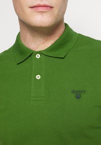 Polo shirt vert en tissu texturé. Présente un col classique, deux boutons et un petit logo brodé bleu sur la poitrine.
