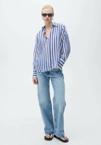 Blau-beige gestreiftes Langarmhemd, geschlossen, kombiniert mit hellblauen geraden Jeans und schwarzen Flip-Flops. Das Modell steht vor einem schlichten Hintergrund.