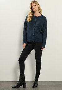Granatowy sweter z dzianiny z okrągłym dekoltem, długimi rękawami i zapięciem na guziki, noszony na czarnych obcisłych dżinsach i botkach na obcasie.