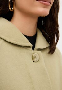 Cappotto verde di lana con colletto, caratterizzato da un grande bottone beige e tessuto strutturato. Una camicia nera è visibile sotto.